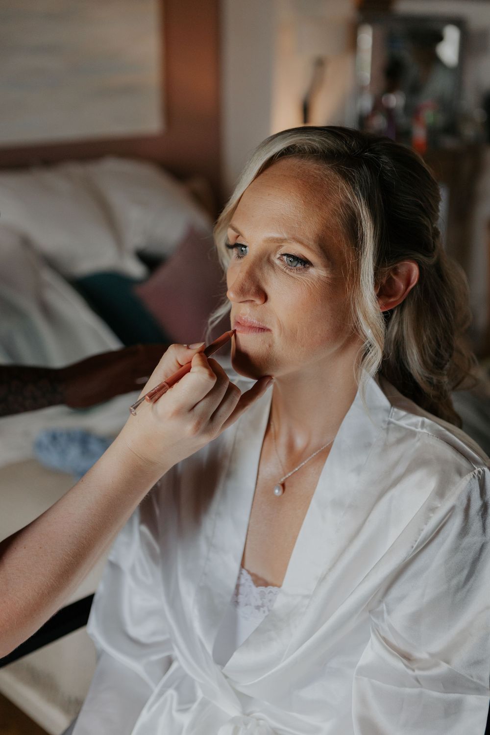 Makeup artist applying lipstick to bride in white robe during wedding preparations.