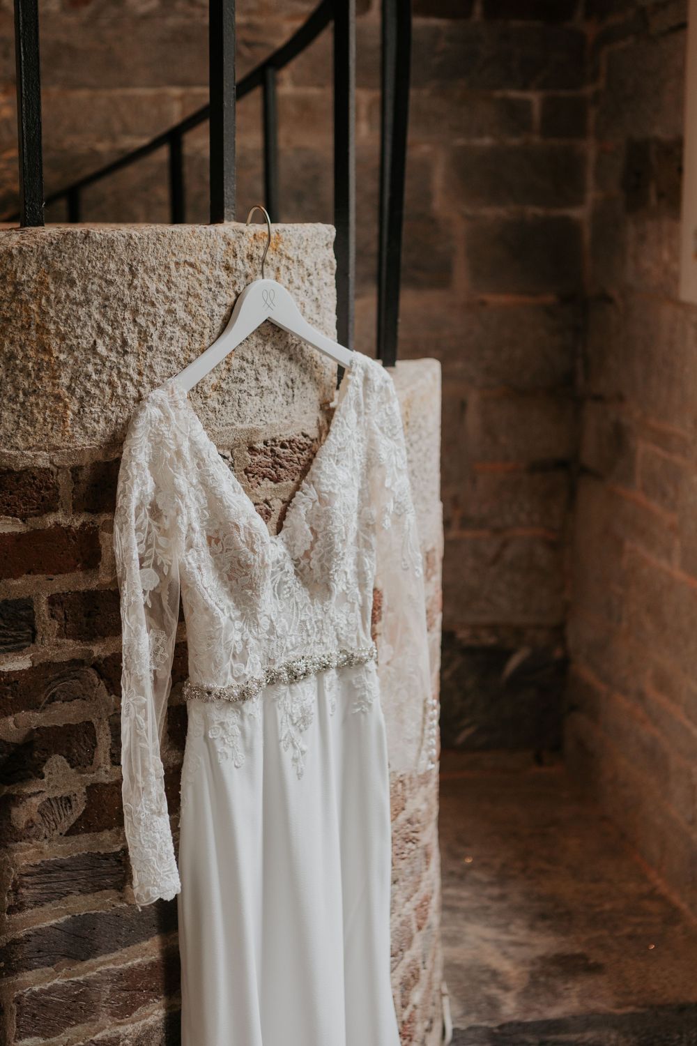 Elegant lace long-sleeve wedding dress hanging on hanger against rustic stone wall.