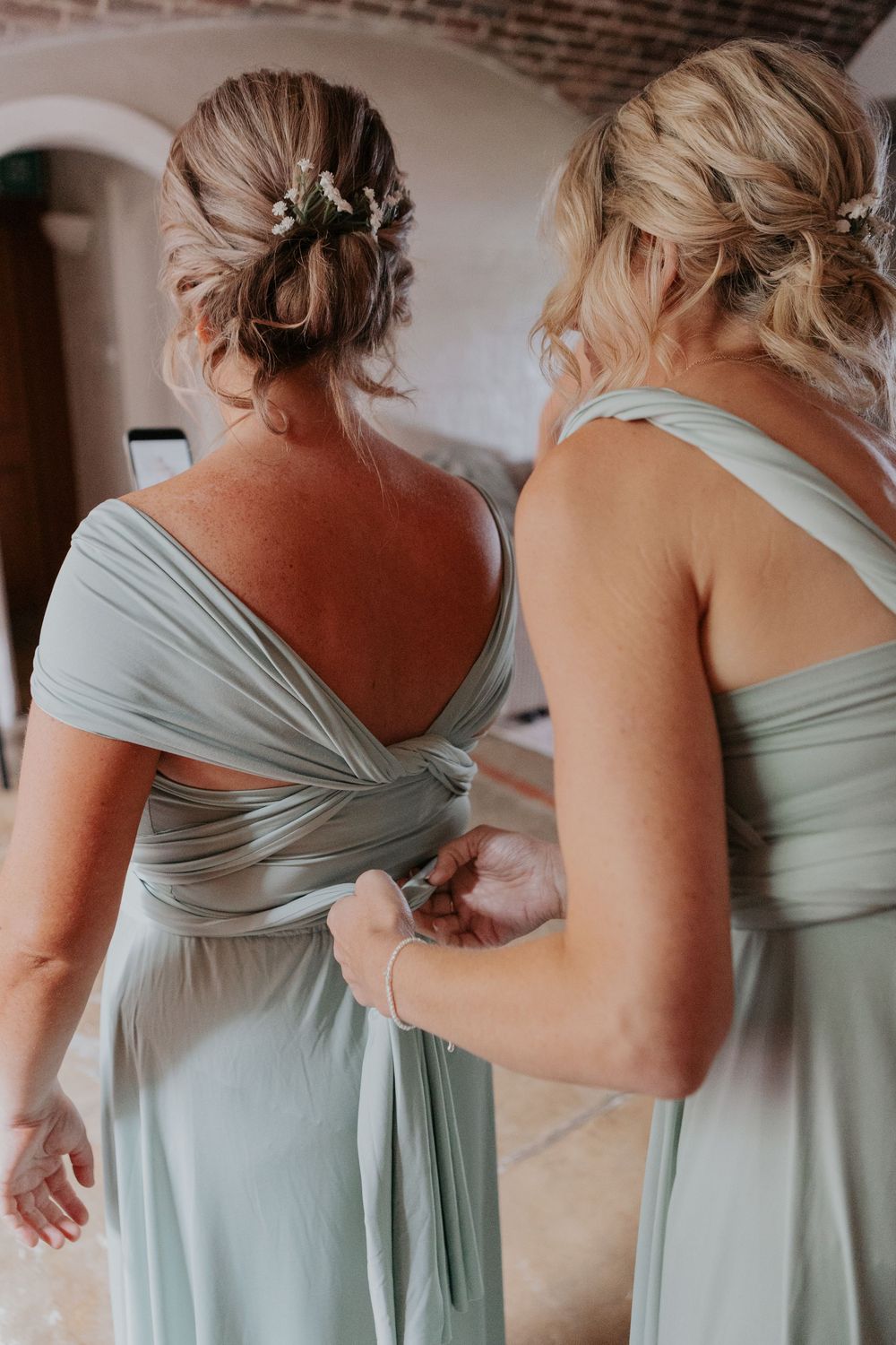 Two bridesmaids in sage green multiway dresses, one fastening the back of the other's gown.