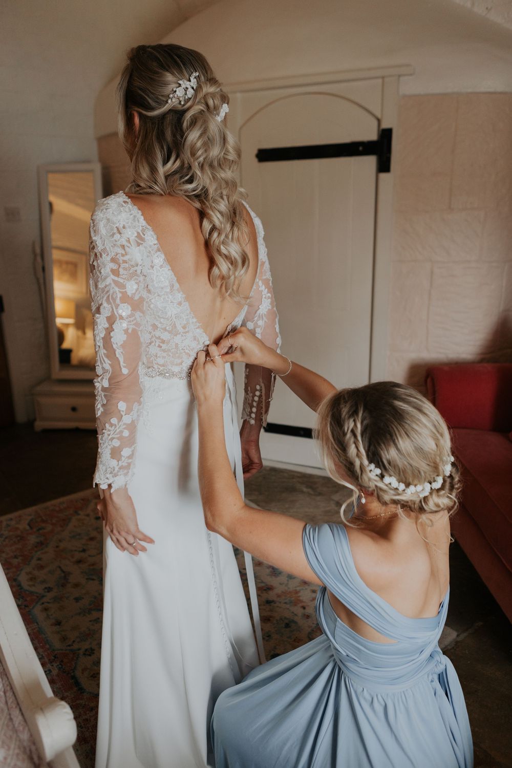 Bridesmaid in blue dress fastening the back of bride's lace wedding gown in a bright room.