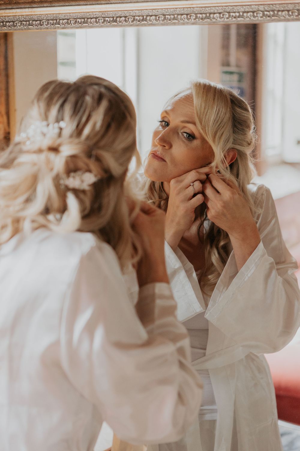 Bride putting on earrings while looking in an ornate mirror during wedding preparations.