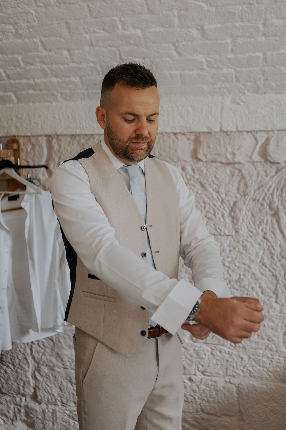 Groom in beige vest and white shirt adjusting cufflinks before wedding ceremony.