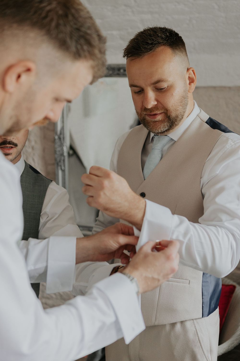 Groomsman helping groom fasten cufflinks while getting ready for wedding.
