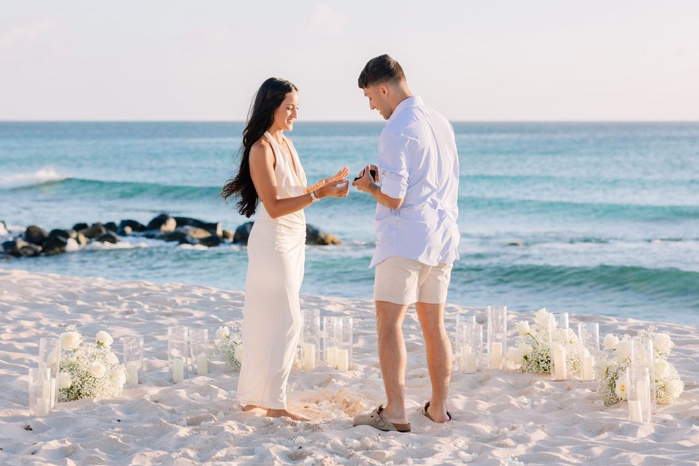 Couple exchanging rings during a romantic beach proposal surrounded by white floral arrangements and candles at sunset.