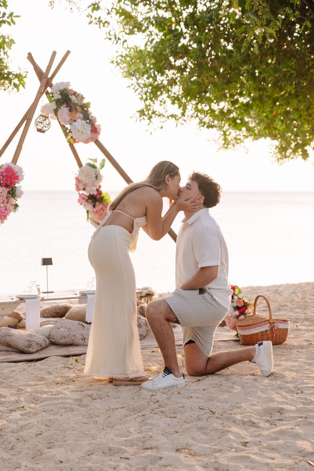 Man proposes to woman on beach with floral arch backdrop at sunset, romantic seaside engagement moment.