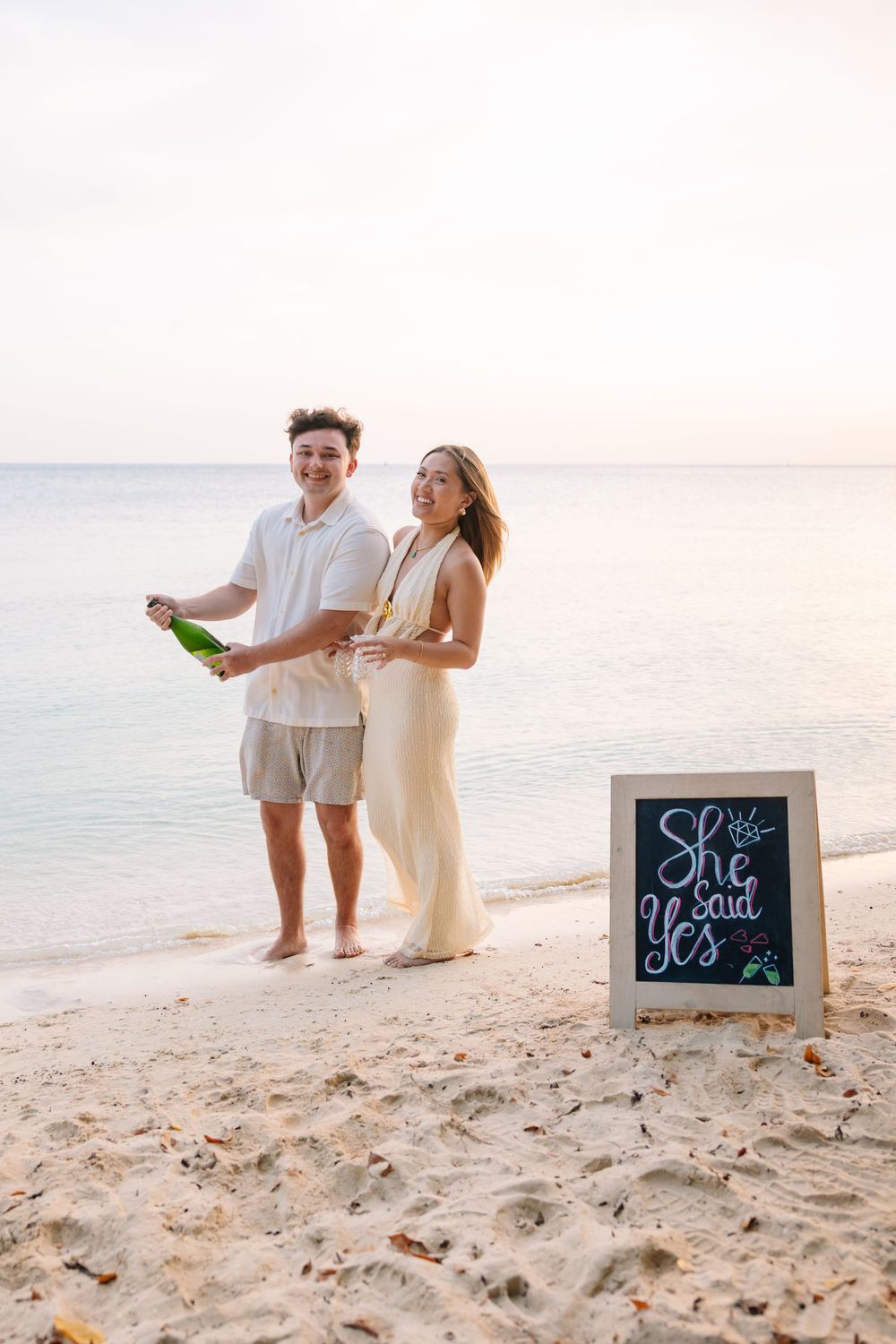 Happy couple celebrating engagement on a beach with champagne and a She Said Yes chalkboard sign at sunset.