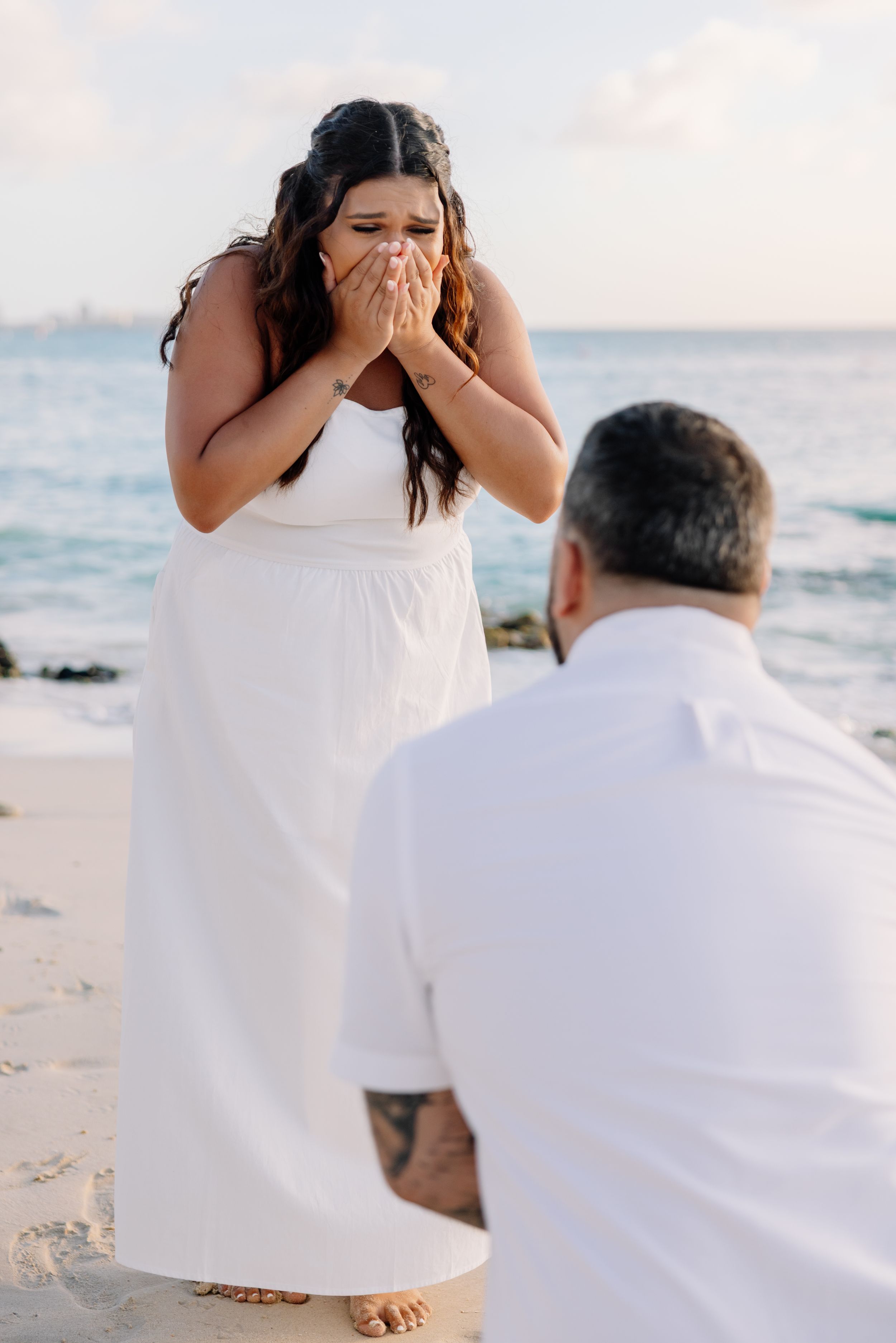 A romantic beach proposal scene showing someone kneeling in white clothing while another person reacts emotionally at sunset.