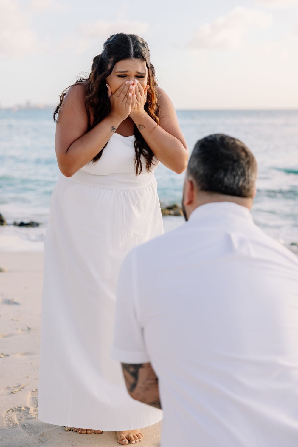 A romantic beach proposal scene showing someone kneeling in white clothing while another person reacts emotionally at sunset.