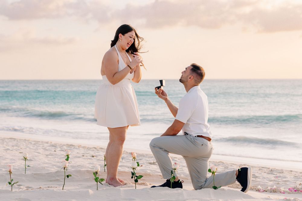 Man proposes to woman on a tropical beach at sunset, kneeling with a ring box while roses are planted in the sand.