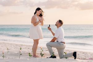 Man proposes to woman on a tropical beach at sunset, kneeling with a ring box while roses are planted in the sand.