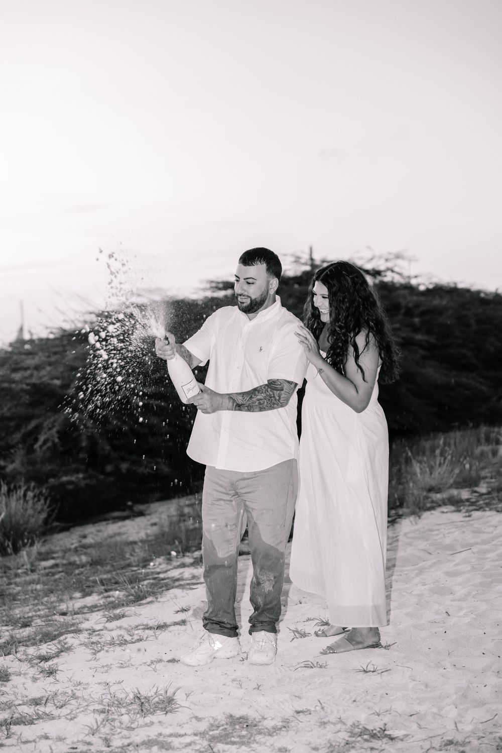 A couple in white clothing playfully throws sand at each other on a beach in a black and white photograph.