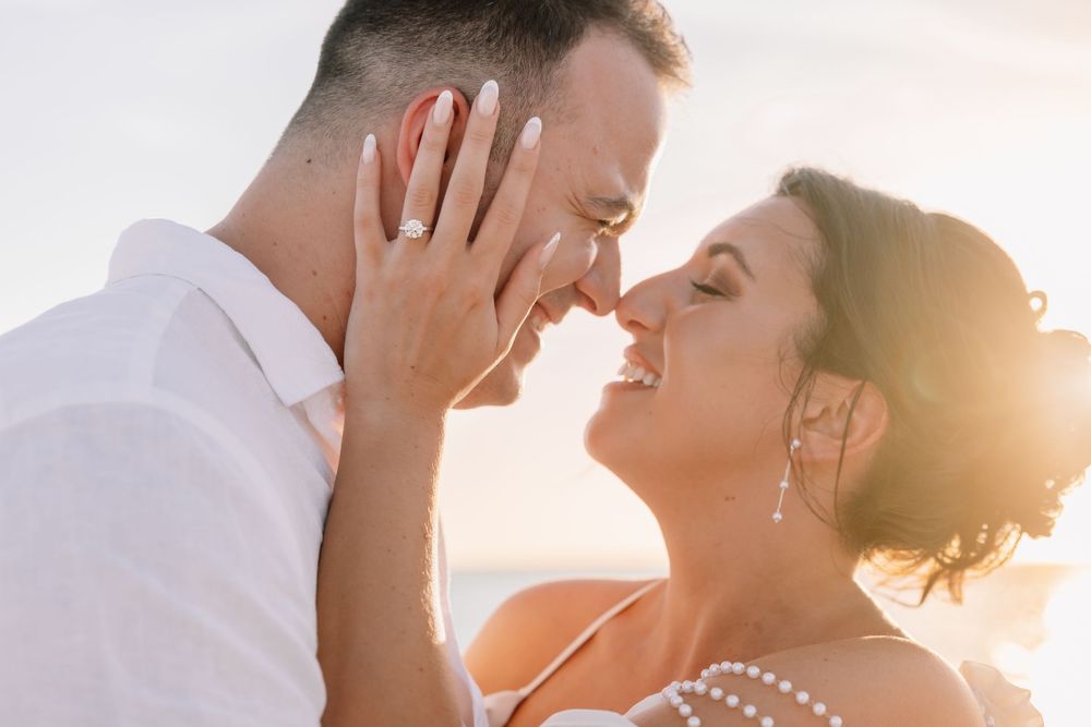 Romantic couple in wedding attire sharing a tender moment at sunset, bride smiling with engagement ring visible.