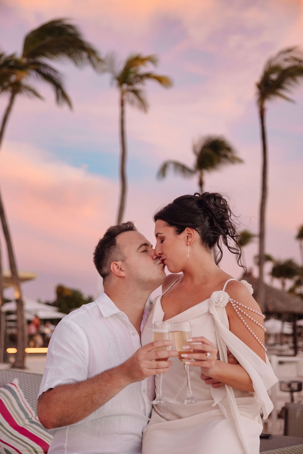 Couple kissing at sunset on tropical beach, holding champagne glasses with palm trees and pink sky backdrop.