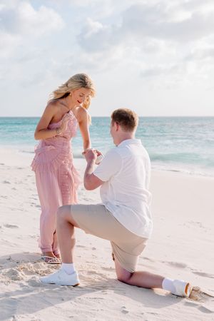 Man proposes on one knee to woman in pink dress on a white sand beach with turquoise ocean backdrop.