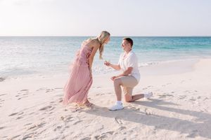 Man proposes to woman on a sandy beach with turquoise ocean waves, she wears a pink maxi dress.