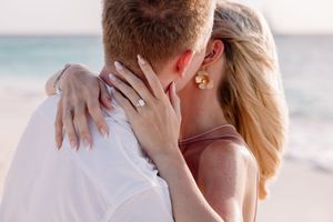 Couple sharing a romantic beach kiss, woman displaying diamond engagement ring, wearing pink dress and gold earrings.
