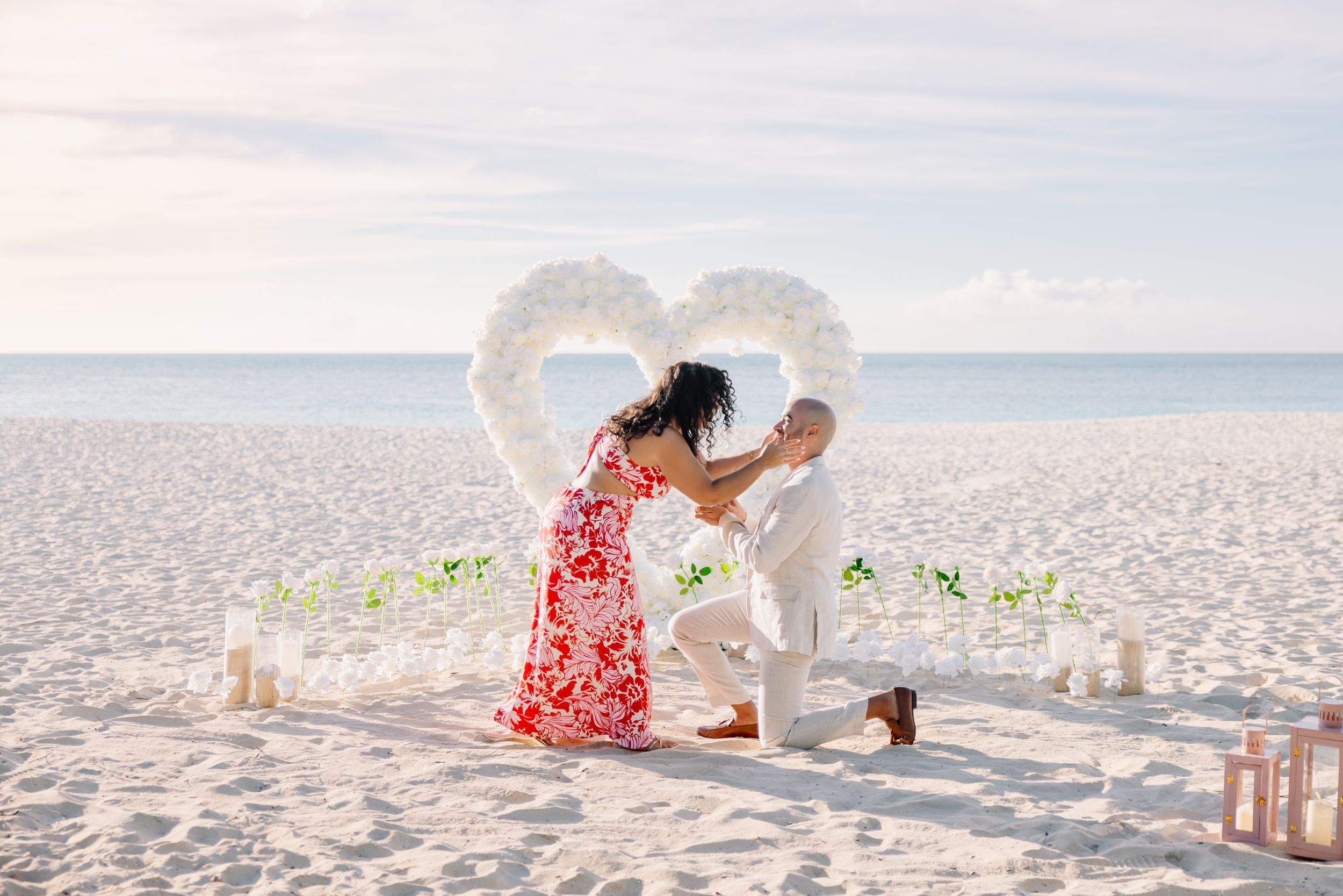 Beach proposal setup with heart arch, candles and couple in romantic moment.
