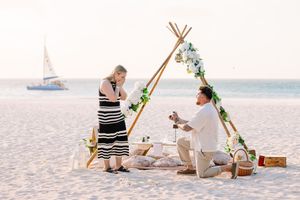 Man proposes to woman on a sandy beach at sunset near a decorated teepee arch with flowers and a sailboat in the background.