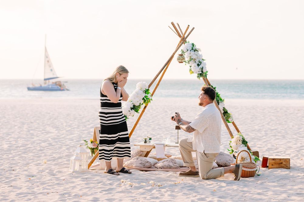 Man proposes to woman on a sandy beach at sunset near a decorated teepee arch with flowers and a sailboat in the background.