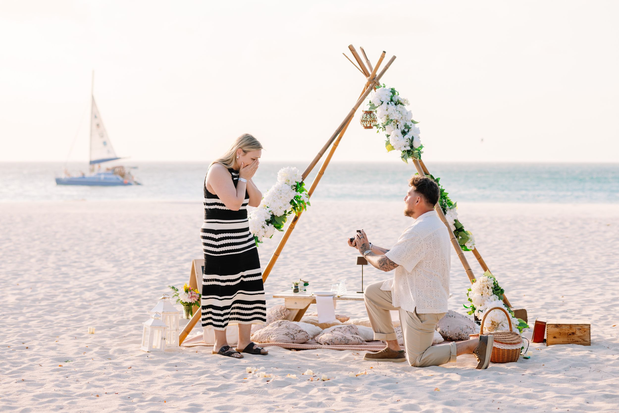 Man proposes to woman on a sandy beach at sunset near a decorated teepee arch with flowers and a sailboat in the background.