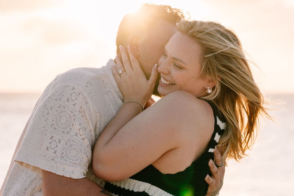 Couple embracing romantically on a beach at golden sunset, woman smiling joyfully while man holds her close.