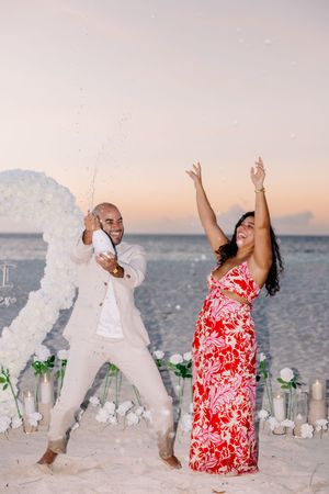A couple in white and red attire dances joyfully on a beach at sunset.