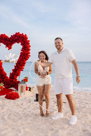 Couple celebrating beach proposal with red rose heart backdrop and “Will you marry me” sign by the ocean.