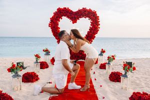 Romantic beach proposal with red rose heart arch, couple kissing on red carpet surrounded by roses and candles at sunset.