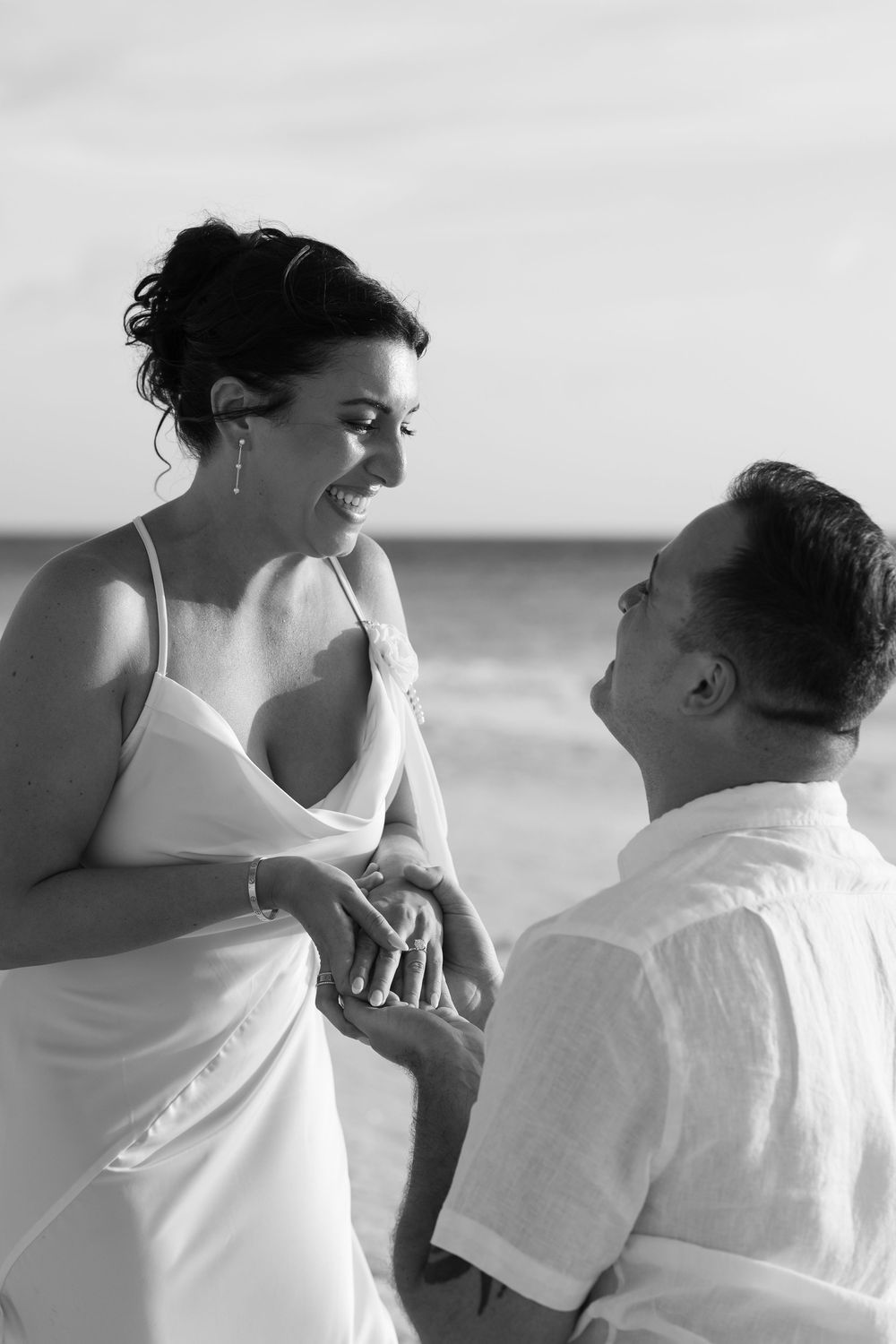 Black and white beach wedding photo of groom kneeling before laughing bride, exchanging rings by the ocean.