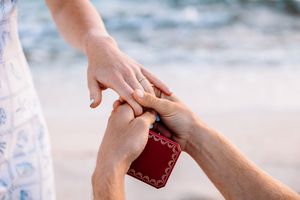 Close-up shot of hands exchanging rings on a sandy beach with ocean waves in the background.