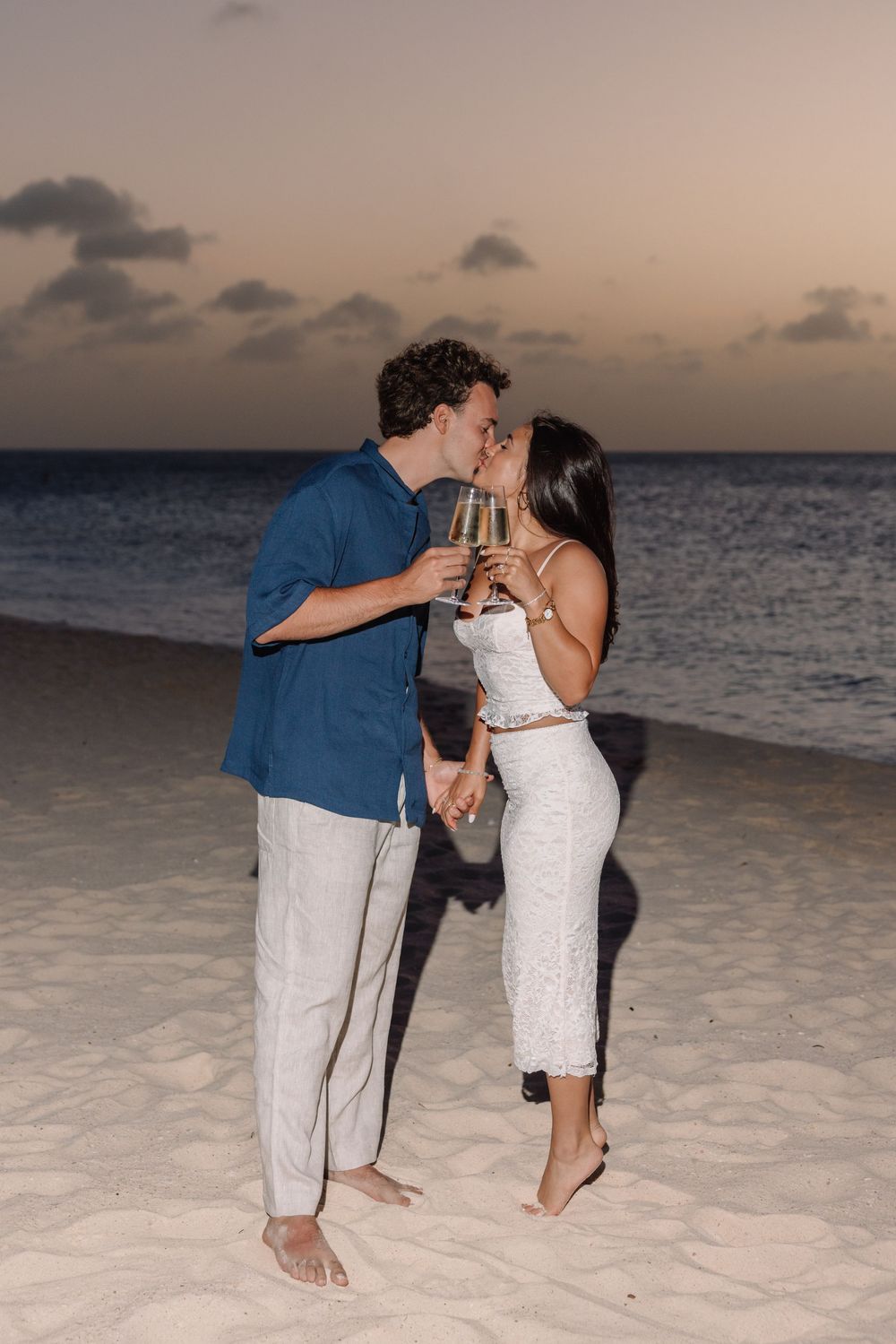 Couple kissing on beach at sunset, holding champagne glasses, romantic evening celebration by the ocean.