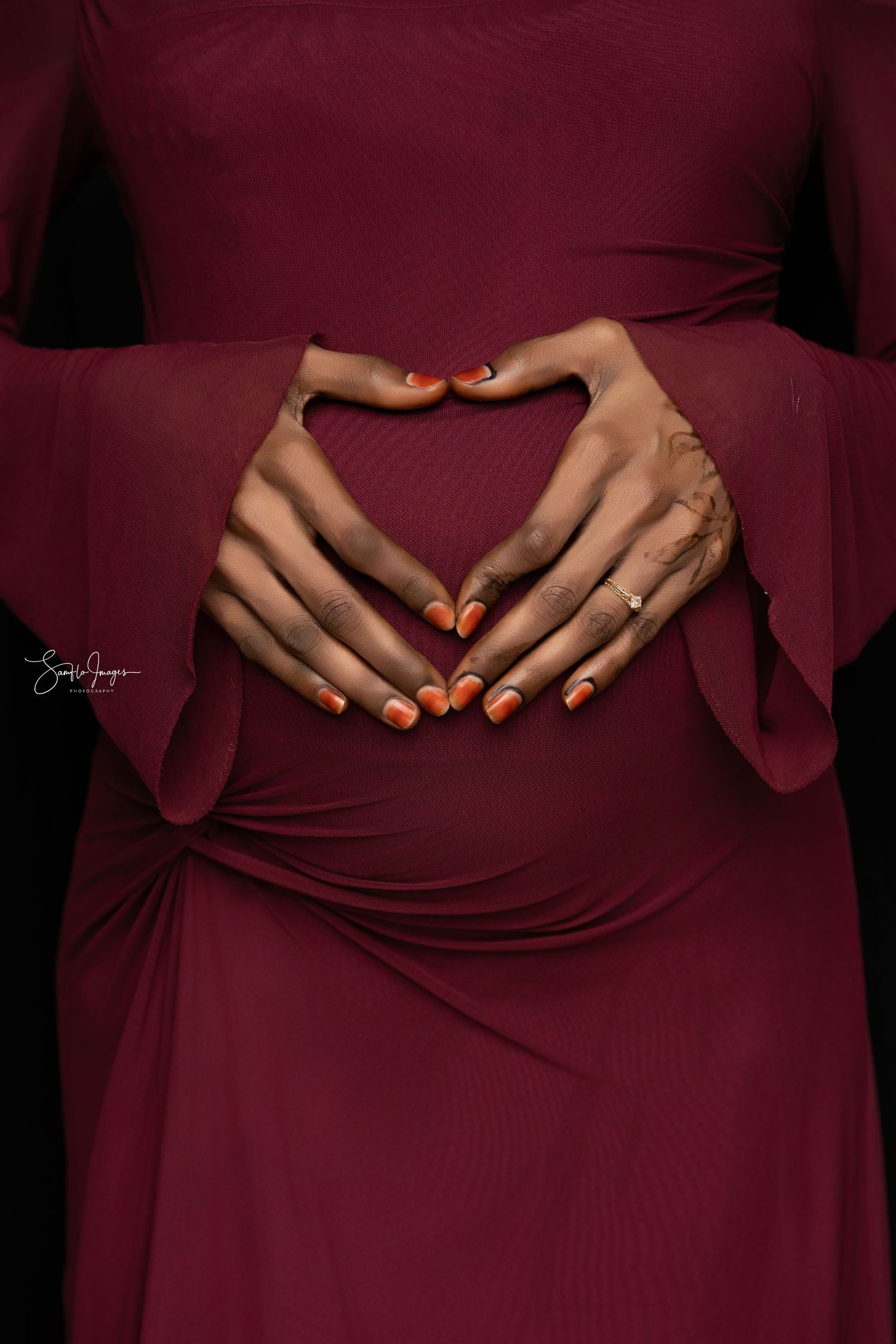 Hands with orange manicured nails form a heart shape against a burgundy dress with bell sleeves.