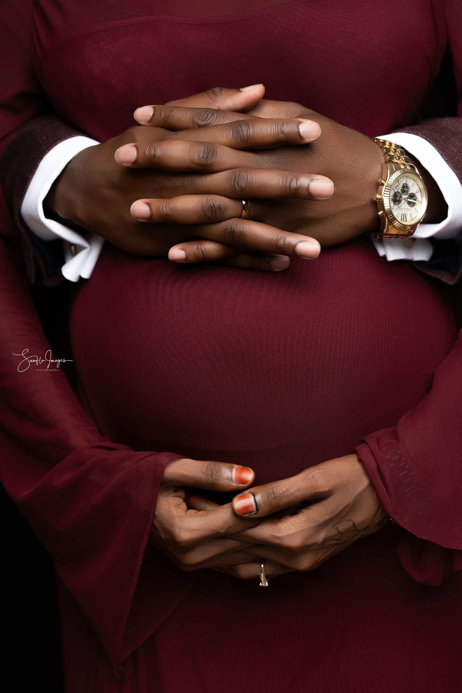 Clasped hands with a gold watch and white cuffs rest against a deep red formal outfit.