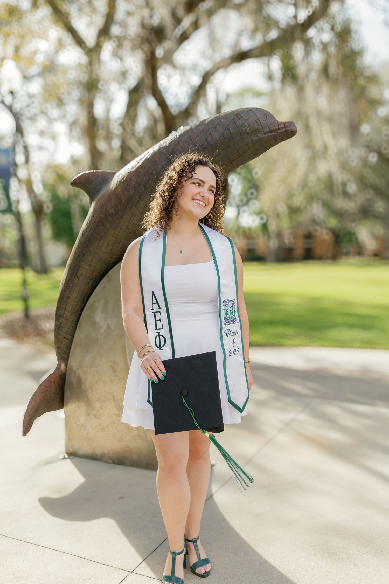 A curly-haired college graduate poses proudly in a white dress with an AEPhi stole beside a dolphin statue on campus.