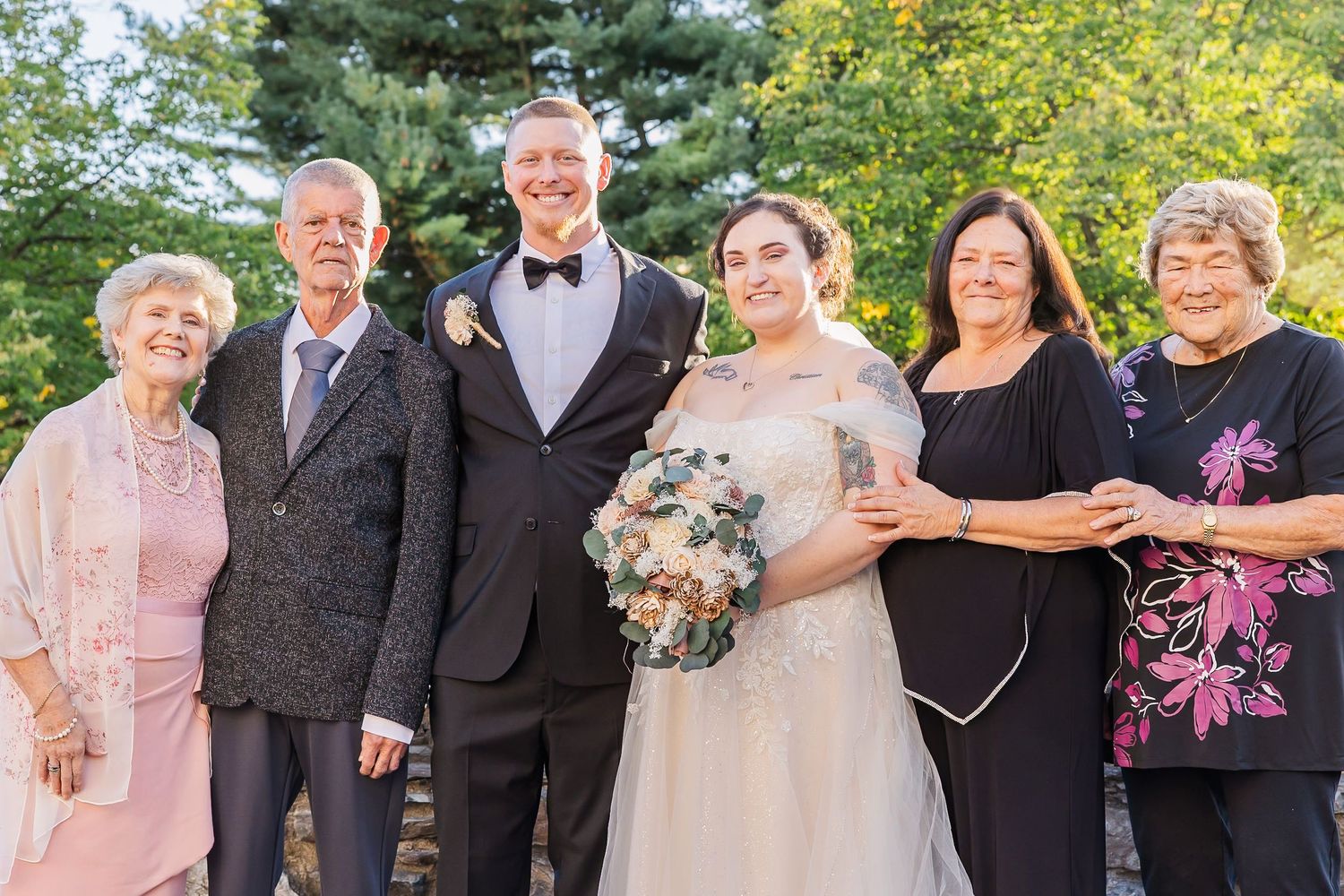 A group of wedding guests pose together outdoors in formal attire with yellow flowers and greenery in the background.