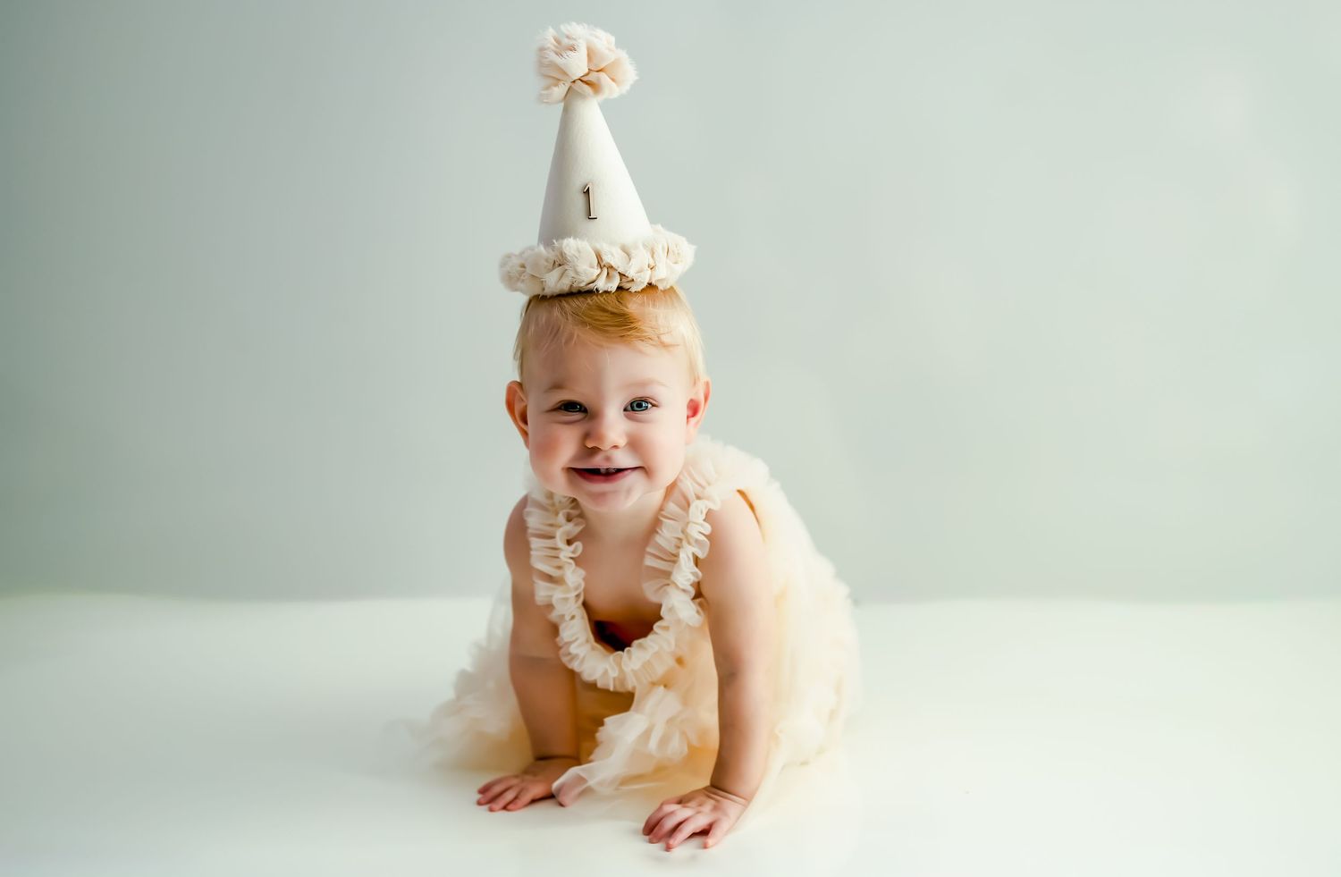 A baby in a white dress and crown sits next to a smash cake during their first birthday photoshoot.