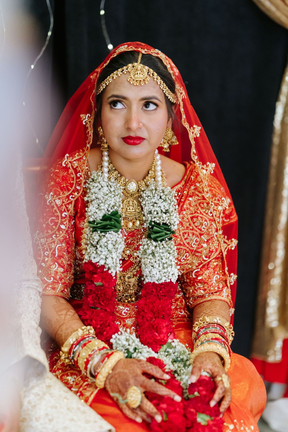 A traditional Indian bride wearing a red and orange bridal sari adorned with floral garlands and ornate gold jewelry.