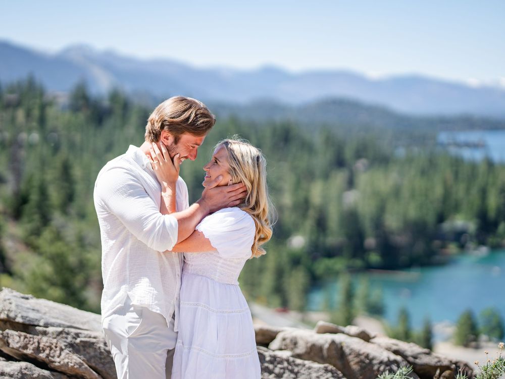 A romantic couple in white outfits embrace on a mountain overlook with a turquoise lake and forest backdrop.
