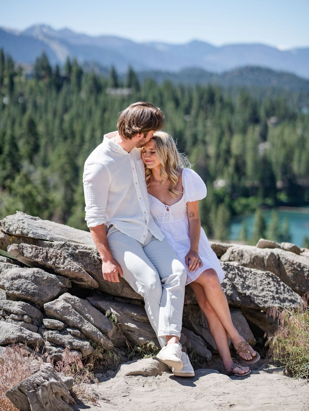 Couple in white outfits sharing a romantic moment on rocky overlook with mountain lake and forest views.