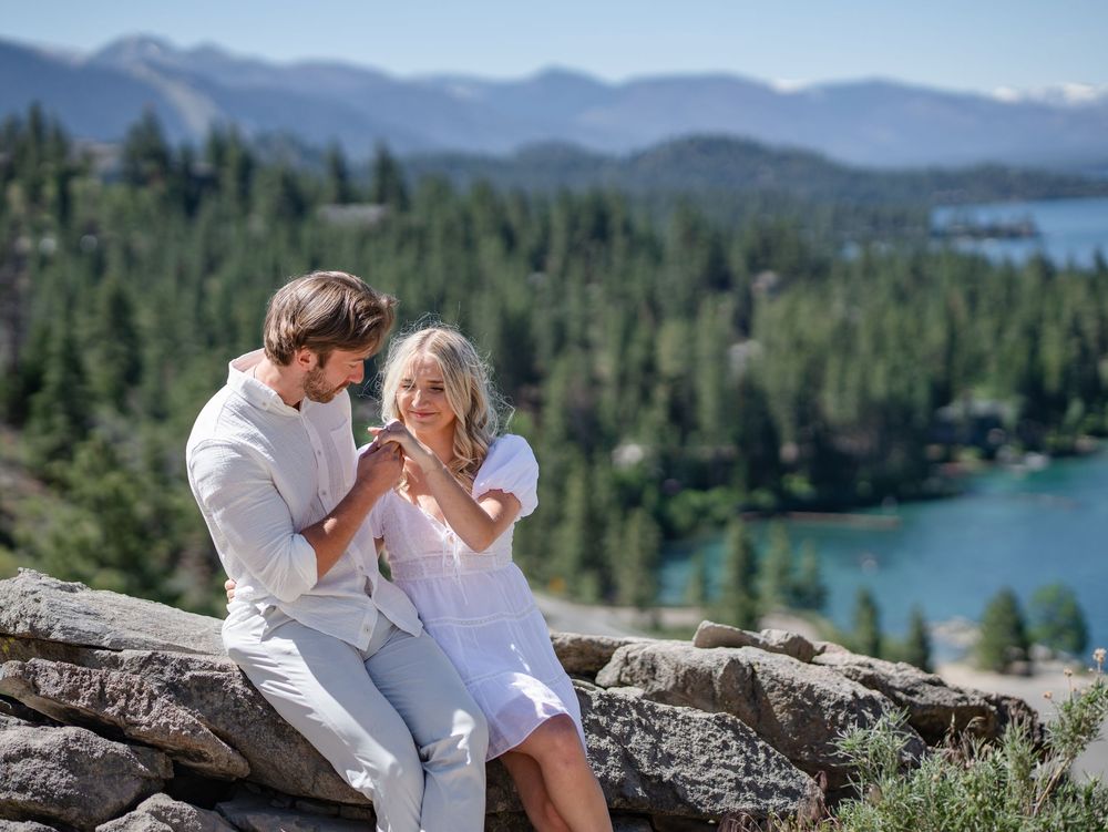 Intimate couple dressed in white sitting on rocks overlooking scenic mountain lake landscape.