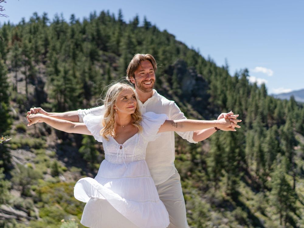 Couple in white wedding attire dances joyfully on a mountaintop with evergreen forest and mountains in the background.