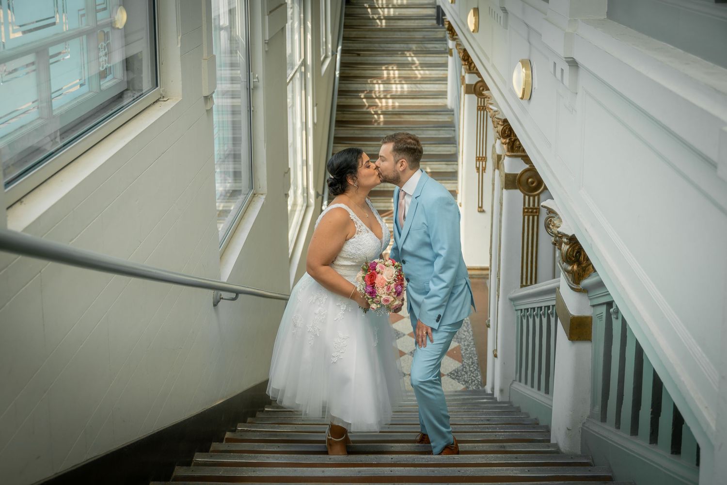 Ein formell gekleidetes Brautpaar posiert vor einem prächtigen gelben Herrenhaus mit weißen Verzierungen an ihrem Hochzeitstag.