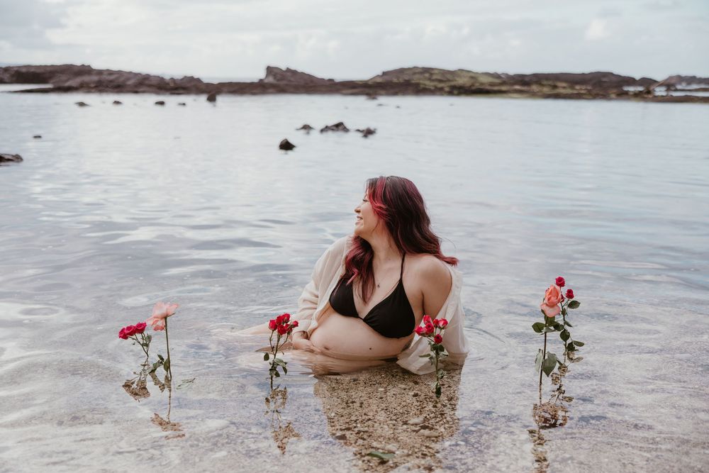 A woman in a black swimsuit poses gracefully in shallow ocean water with red roses creating an artistic beach scene.