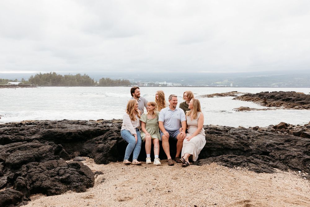 A group sits together on volcanic rocks at a scenic beach overlook in Hawaii with waves crashing in the background.
