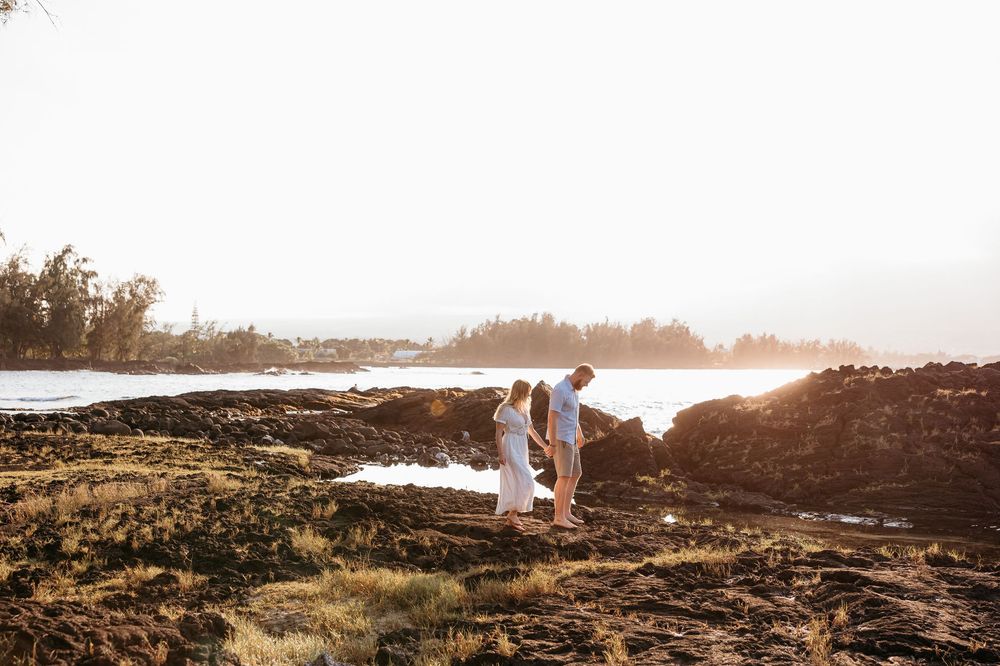 Couple walks along scenic rocky coastline at sunset with ocean waves crashing against cliffs in background.