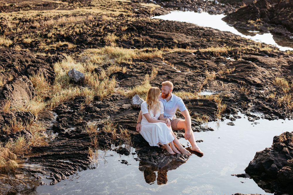 A couple sits together on rocky cliffs overlooking a scenic coastal landscape at sunset.