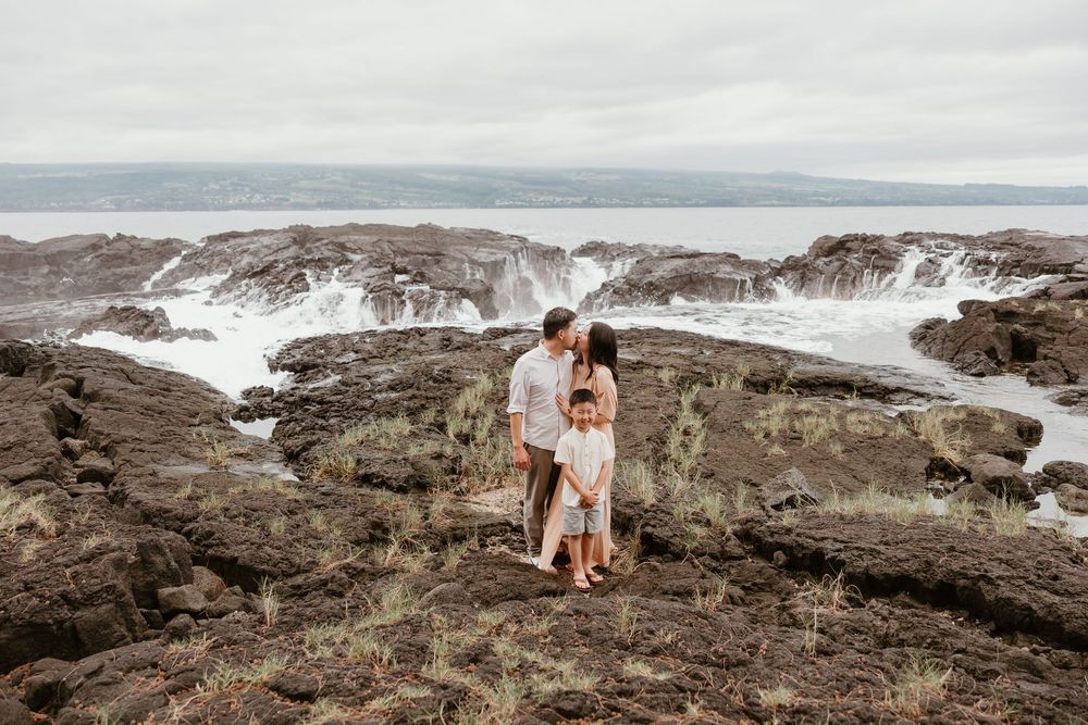 A couple stands together on rugged coastal cliffs overlooking dramatic ocean waves and rocky shoreline at sunset.