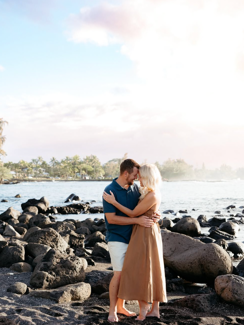 A couple embraces on rocky beach shoreline with palm trees and ocean waves in Hawaii at sunset.