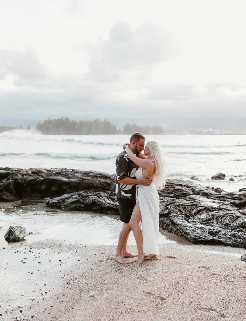A couple embraces lovingly on a rocky beach with crashing waves and dramatic cloudy skies in the background.