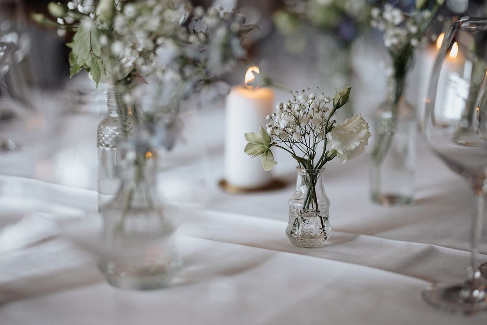 Small glass bud vases with delicate white flowers create an elegant table setting at a wedding reception.